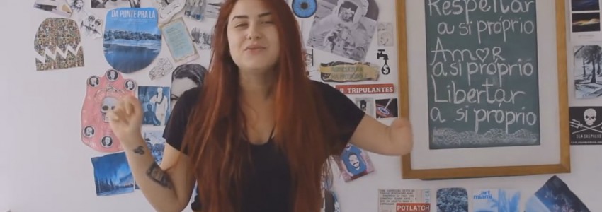 Em um quarto com várias fotos, desenhos e textos na parede, uma jovem está de pé. Ela é branca de cabelos avermelhados e olhos claros, e fala gesticulando. A jovem não tem parte do braço esquerdo.