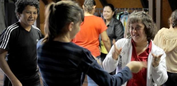 Em uma sala fechada, várias pessoas jovens estão em duplas, em um exercício de teatro. Uma jovem com Síndrome de Down sorri olhando na direção de outra jovem, durante o exercício