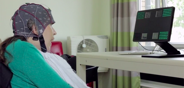 Uma mulher com mobilidade reduzida, sentada em uma cadeira, com uma touca cheia de eletrodos; ela olha na direção de um monitor de computador
