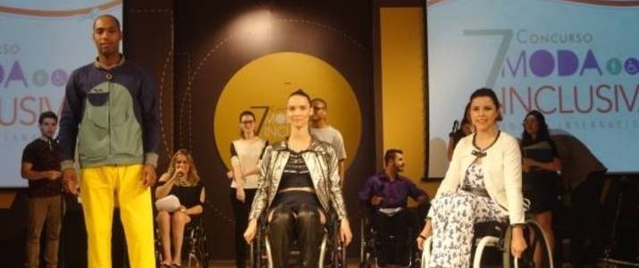 Foto do desfile com roupas inclusivas; três modelos estão em uma passarela. O primeiro modelo é um homem negro e jovem, à esquerda; as duas outras modelos, mulheres brancas e jovens, estão em cadeira de rodas