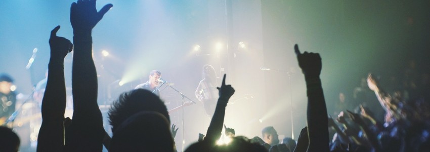 Foto de um show com destaque da multidão com as mãos para cima assistindo banda tocar em um palco.