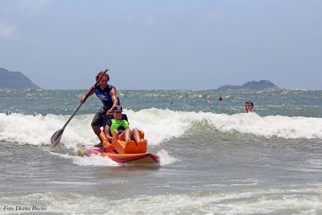 Foto em uma praia em um dia ensolarado. Mara Gabrilli está em uma prancha de surf adaptada para ela. Na mesma prancha está um homem que oferece apoio e segura um remo. 