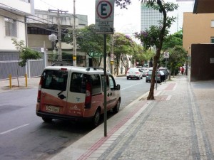 Calçada com piso tátil interrompido por árvores e placa de sinalização