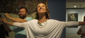 Mulher de braços abertos sorri olhando para cima enquanto um homem toca suas mãos