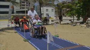 Em uma praia, um tatame de borracha forma o caminho da areia até o mar; há um jovem cadeirante sendo guiado nesse percurso por outro homem, mais velho.