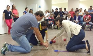 Um homem e duas mulheres estão mexendo com algumas bolinhas que estão pelo chão. Ao fundo, uma plateia acompanha a atividade.