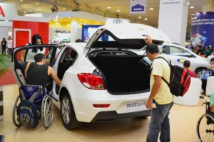 Um homem de amarelo e um rapaz na cadeira de rodas observam um veículo branco adaptado, que está em um estande da Mobility & Show
