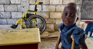 m menino de aproximadamente 6 anos, negro, amparado por muletas, olha para a câmera com uma expressão triste