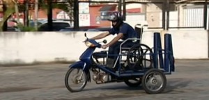 Rapaz pilota a moto adaptada. Ela possui três rodas, como um triciclo, com espaço para a cadeira de rodas.