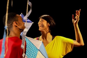 Foto de um espetáculo de dança; uma jovem mulher, com uma blusa amarela, sorri com os braços arqueados, enquanto um outro homem interage com ela; há pedaços coloridos de tecido descendo do teto