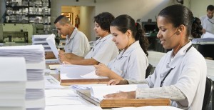 Em um ambiente amplo, fechado e bem iluminado, três mulheres e um homem estão sentados em bancos, manuseando papéis e livros em braile dispostos em uma bancada