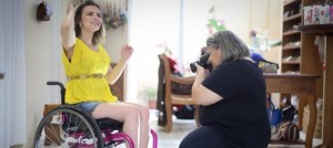 Foto de uma jovem cadeirante sorrindo e posando para a fotógrafa que a clica em uma sala ampla e iluminada