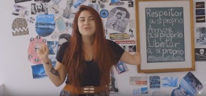 Em um quarto com várias fotos, desenhos e textos na parede, uma jovem está de pé. Ela é branca de cabelos avermelhados e olhos claros, e fala gesticulando. A jovem não tem parte do braço esquerdo.