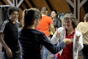 Em uma sala fechada, várias pessoas jovens estão em duplas, em um exercício de teatro. Uma jovem com Síndrome de Down sorri olhando na direção de outra jovem, durante o exercício