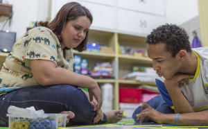 Em uma biblioteca escolar, uma jovem professora, de cabelos curtos, acompanha um aluno mais velho, com deficiência, observando um tablet