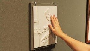 Foto de uma obra em alto relevo sendo tateada por uma mão feminina; a imagem tátil é de um senhor sentado em frente a uma mesa