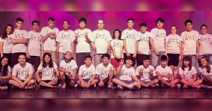 Em um palco de teatro, os atores com espectro autista posam para a foto, disposto lado a lado e sentados no chão