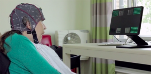 Uma mulher com mobilidade reduzida, sentada em uma cadeira, com uma touca cheia de eletrodos; ela olha na direção de um monitor de computador