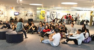 Foto de uma das salas do Memorial da Inclusão; a sala é ampla, com colunas e obras penduradas nas paredes; um grupo de pessoas de diversas idades está sentada no chão, enquanto uma mulher, sentada em um puff, conversa com eles