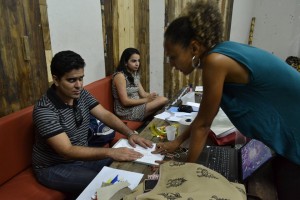 Foto de uma sala de aula, com uma longa bancada. Uma mulher está de pé e orienta um dos alunos, um homem cego que está sentado. Ao lado dele, há uma jovem.