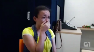 Em ambiente interno, mulher está sentada e chora emocionada, com a mão direita cobrindo a boca.