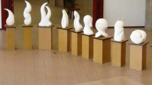 Foto traz nove esculturas da exposição Movimento em Branco. Apoiadas sobre totens, elas são pequenas, brancas e possuem formas abstratas.