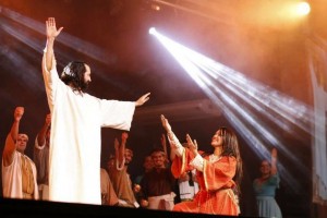 Cena do musical traz um ator caracterizado como Jesus. Ele tem um dos braços erguidos e abençoa, com o outro, uma mulher que está agachada e sorri para ele com os mãos em sinal de pedido.