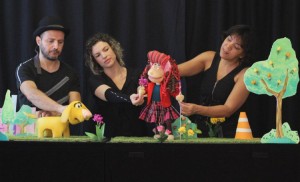 Foto do espetáculo. Em um palco, três pessoas manipulam fantoches de uma menina e um cachorro