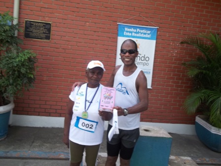 Foto da senhora Dona Maria de Lourdes ao lado do seu filho Rogério no pódio. Os dois estão posando para a foto, sorrindo. Ela usa uma medalha e roupa de corrida e segura um troféu com Rogério.