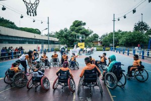 Foto do time de basquete de cadeira de rodas, na quadra. Dispostos em um círculo, os atletas olham para uma mulher que está de pé. Mais ao fundo, outro time está disposto em um círculo.