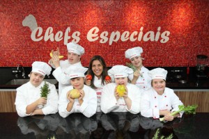 Foto de uma cozinha, com um grupo de seis alunos com Síndrome de Down, de aproximadamente 10 anos de idade, e uma jovem professora ao centro. Todos posam para a foto segurando ingredientes e vestem domãs e chapéus de cozinheiros