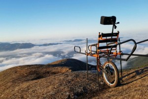 Foto em plano aberto de cadeira de rodas adaptada, com apenas uma roda e barras para que ela possa ser carregada, no topo de uma montanha