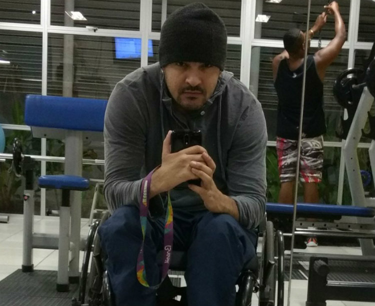 Foto de um homem moreno cadeirante, de 29 anos. Ele é malhado, usa touca e cavanhaque e faz uma selfie na frente do espelho, com o celular na mão