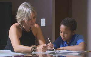 Foto de uma mulher loira ajudando um garoto negro com o dever de casa.