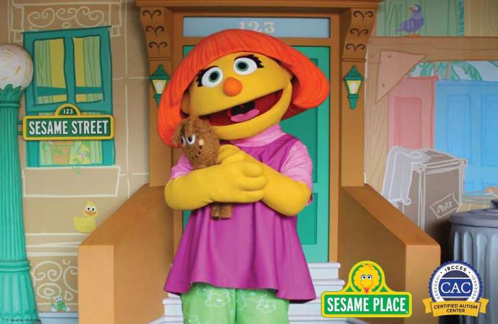Foto da personagem Julia, da Vila Sésamo, segurando um ursinho de pelúcia. Ela tem uma aparência lúdica, com uma cabeça grande e amarela, nariz e cabelos laranja