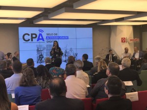 Em um auditório fechado, uma mulher se apresenta ao público. Ela é alta, morena e está de pé. Na tela ao fundo, há o texto "CPA e Selo de Acessibilidade Digital"
