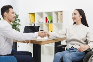 Um homem aperta a mão de uma mulher cadeirante. Os dois sorriem.