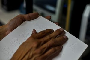 Foto em close de duas mãos deslizando sobre uma folha de papel com texto em braile.