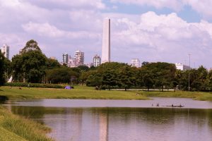 Foto paisagem do Parque do Ibirapuera. O céu está azul e com nuvens brancas, e há muitas árvores, grama e um lago no centro da imagem. Ao fundo, o Obelisco e Monumento Mausoléu ao Soldado Constitucionalista de 32.