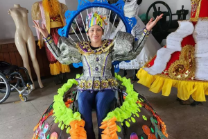 foto de mulher sentada em uma cadeira de rodas. Ela veste uma fantasia para desfile de carnaval, está com os braços levantados e sorri. Ao fundo, há manequins com outras fantasias.