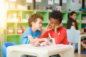 Foto de duas crianças estudando e interagindo com um livro em uma sala de aula.