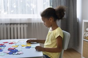 Foto de uma menina, sentada em uma mesa, montando um quebra-cabeça colorido. Ao fundo, há uma janela e cortinas.