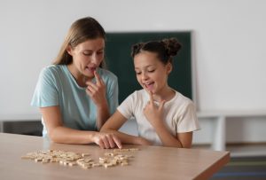 Foto de uma psicóloga auxiliando uma criança na terapia de fala. As duas estão com o dedo indicador apontando para a boca enquanto interagem com peças de um quebra cabeça que está sobre uma mesa.