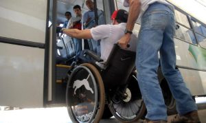 Foto de uma pessoa cadeirante tendo dificuldades ao subir as escadas de um ônibus.