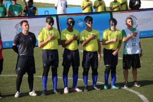 Foto de seis jogadores da seleção brasileira de futebol de cegos em pé no campo de futebol durante o dia.