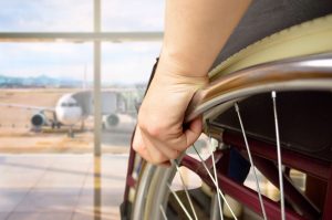 Foto em detalhe de pessoa em uma cadeira de rodas com a mão apoiada na roda. No fundo desfocado, há um avião.