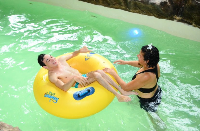 Foto de um homem com deficiência em cima de uma boia amarela ao lado de uma mulher em pé, ambos estão dentro de uma piscina com a água bem verde