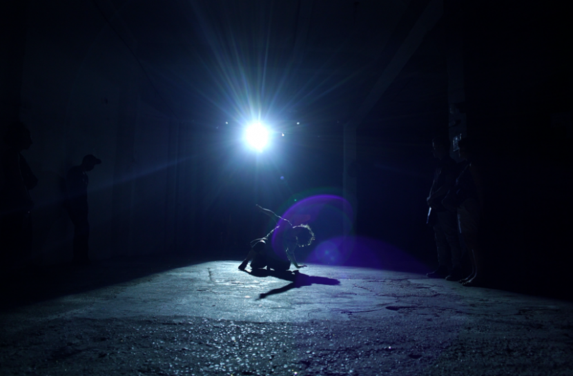 Foto de uma das dançarinas em pose próxima ao chão, com uma das mãos levantada. Ao fundo, uma fonte de luz que ilumina o local. Nas laterais, pessoas assistem em pé.