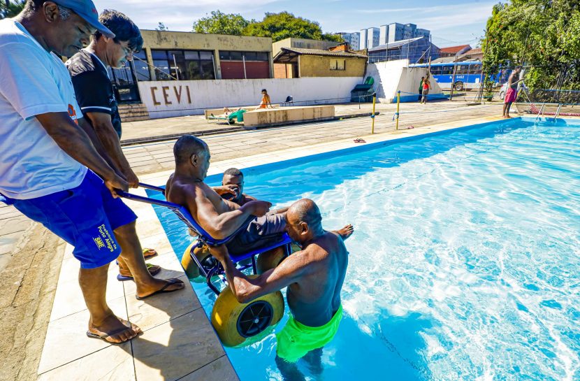 Homem entra na piscina sentado em cadeira anfíbia com auxílio de outras quatro pessoas