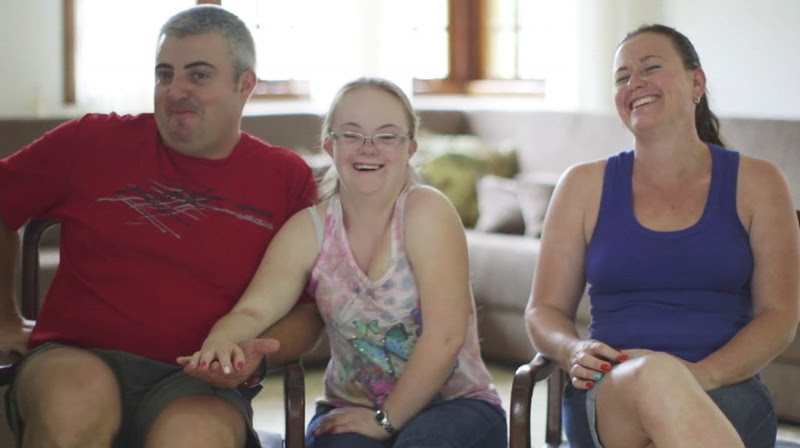 Um homem, uma menina com Síndrome de Down e uma mulher sorriem para a câmera